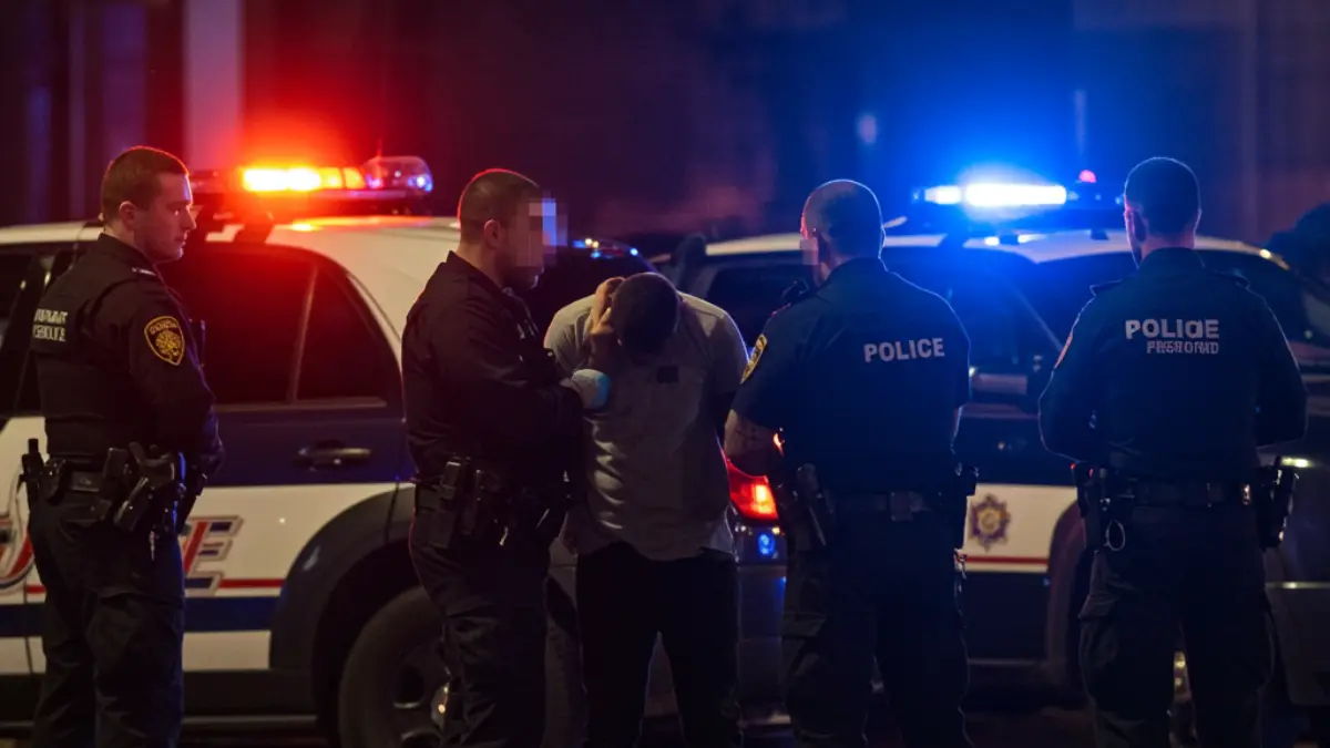 Four police officers detain a man at night beside patrol cars with red and blue emergency lights flashing.