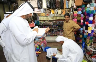 Man in white traditional clothing writes on a clipboard in a crowded market stall filled with handbags and colorful scarves; another man stands nearby watching.