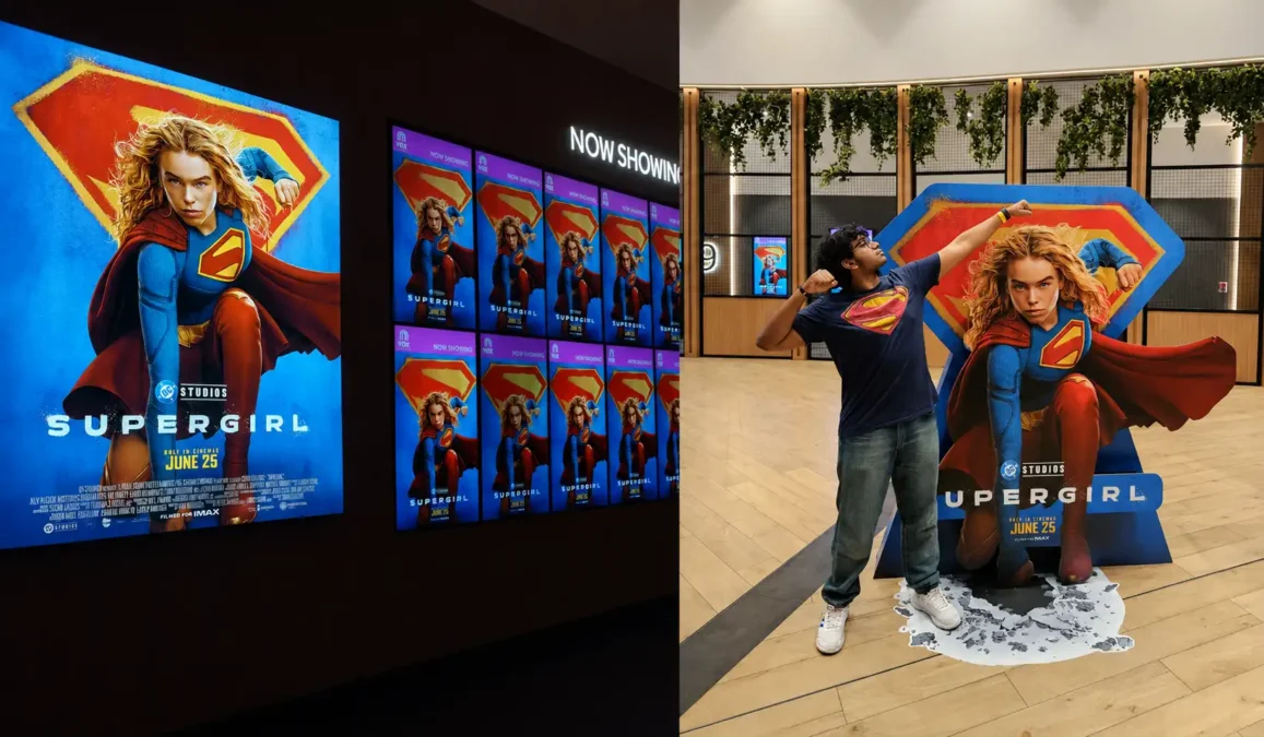 A fan posing like a superhero beside a supergirl movie standee with supergirl cinema posters and promotional screens in the background Times of dubai